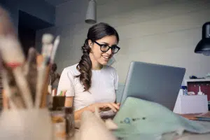 Small business owner woman sitting at desk smiling at laptop with paint brushes and supplies around her.